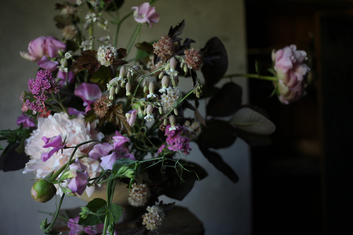 A rustic floral arrangement featuring pinky purple sweet peas and pale pink peonies with dark purple ninebark, photographed in moody lighting.