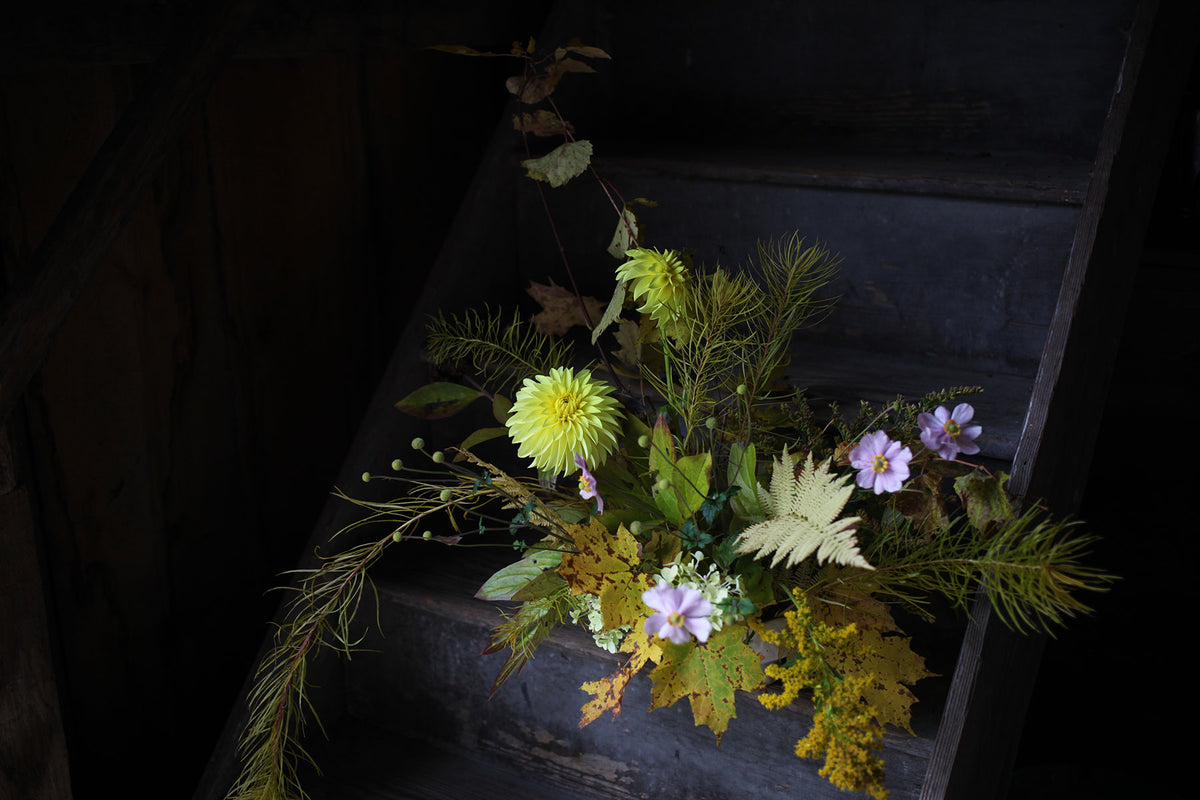 A rustic floral arrangement featuring large yellow dahlias, Japanese anemone, and yellow-tinged foliage, placed on wooden steps in a dimly lit barn.